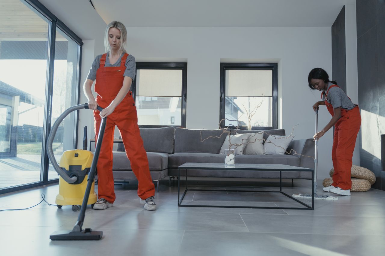 Deux femmes en salopette rouge passent l’aspirateur et balaient un salon moderne.